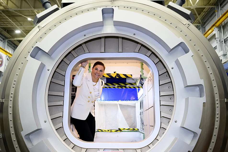 Виктория в делови тъмен панталон и сако стои пред голяма, метална космическа капсула в хангар на НАСА, гледайки с интерес към вътрешността ѝ.