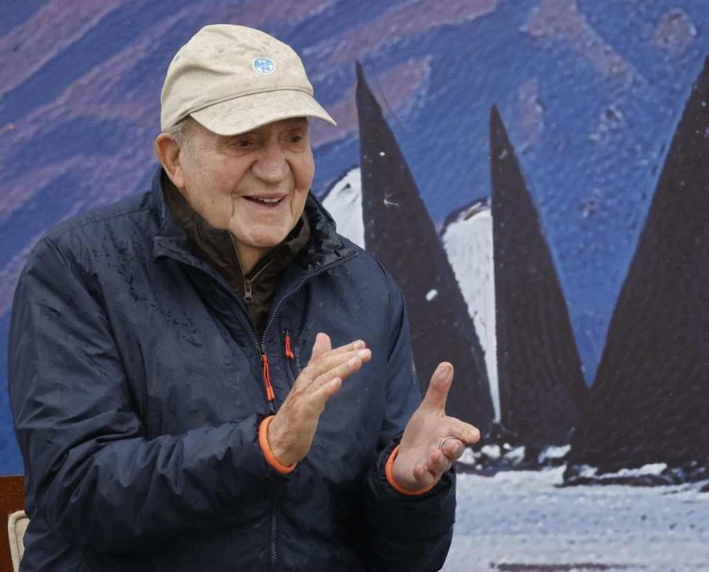 Усмихнатият Хуан Карлос I в тъмносиньо спортно яке и бежова шапка на фона на ветроходни мотиви.