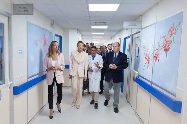 Принцът и принцесата на Монако вървят по модерен болничен коридор, придружени от официални лица и медицински персонал.