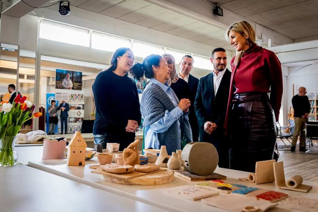 Кралица Максима, заобиколена от доброволци и местни жители, разглежда маса с ръчно изработени глинени фигурки в общностния център „De Bol“.