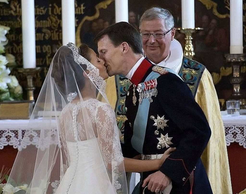 Младоженци се целуват пред олтар пред погледа на правостоящ свещеник.