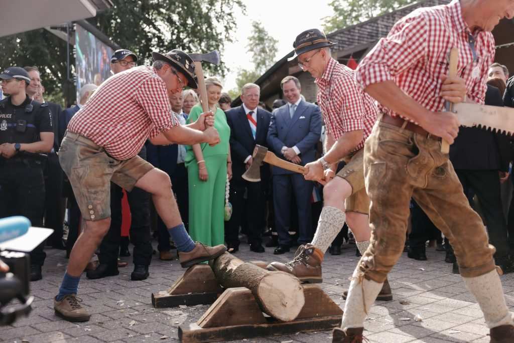 Семейството наблюдава мъже в народни носии, които режат и цепят дървени трупи. 